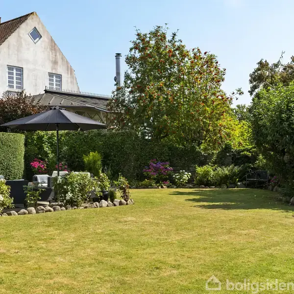 Græsplæne med havemøbler under en parasol, omgivet af velplejede buske og et blomstrende træ. I baggrunden ses et hvidt hus med rødt tag og blå himmel.