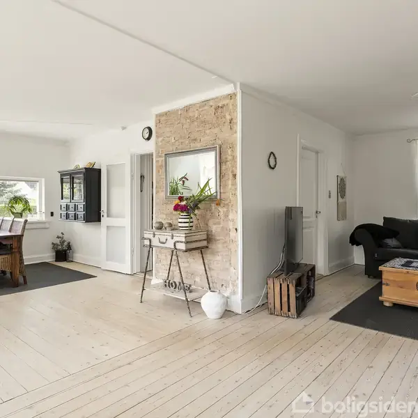 Stue med hvidmalede trægulve, et sofabord foran en mørk sofa og et spisebord ved et vindue. Væg i rå mursten deler rummet. Klar, lys indretning.