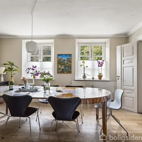 Ovalt spisebord omgivet af stole, står centralt med en vase med blomster og to stearinlys. Rummet er lyst med to vinduer, placeret i et indbydende spisekøkken.