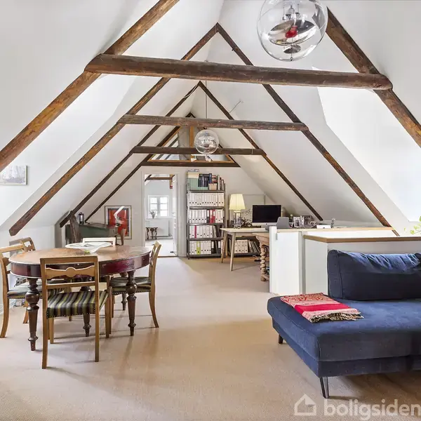 Tagbjælker strækker sig over et rummeligt loftsrum med skrå vægge. En blå sofa og spisebord er placeret centralt. Et skrivebord og reol ses bagerst, omgivet af lys og planter.