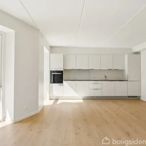 Lyst køkken med hvide skabe og indbygget ovn, set mod en stor have gennem et bredt vindue. Gulvet er lyst træ.
