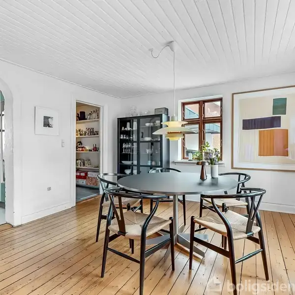 Et rundt spisebord med fire stole står centralt i en lys stue med trægulv. En moderne lampe hænger over bordet. I baggrunden ses et åbent køkken og en reol.