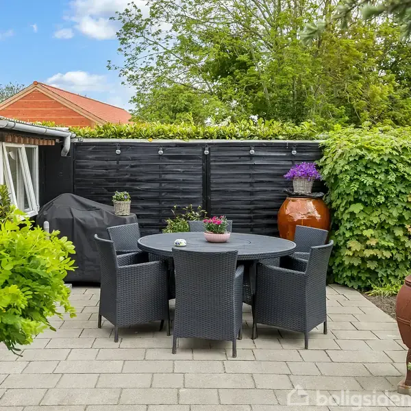 Rundt havebord med fem sorte stole står på flisebelagt terrasse. En grill dækket med sort betræk ses ved siden af, omgivet af grønne planter og træhegn.