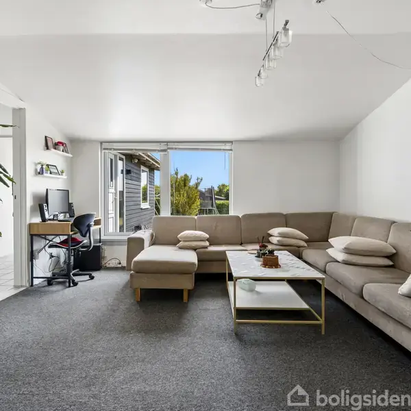 En stue med en stor beige hjørnesofa omkring et sofabord. Til venstre står et skrivebord med stol, og en stor plante. Et vindue viser udsigt til en terrasse.