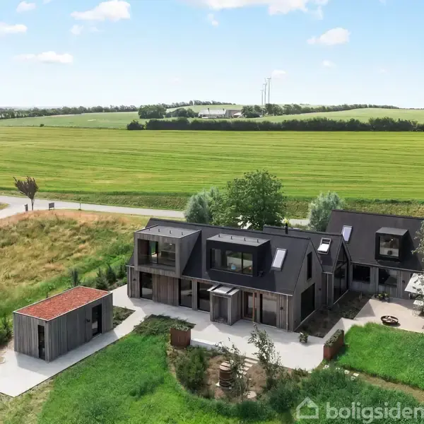 Et moderne, sortmalet hus med store vinduer omgivet af grønne marker under en lys himmel. En mindre bygning står til venstre, mens en grusvej fører til ejendommen.