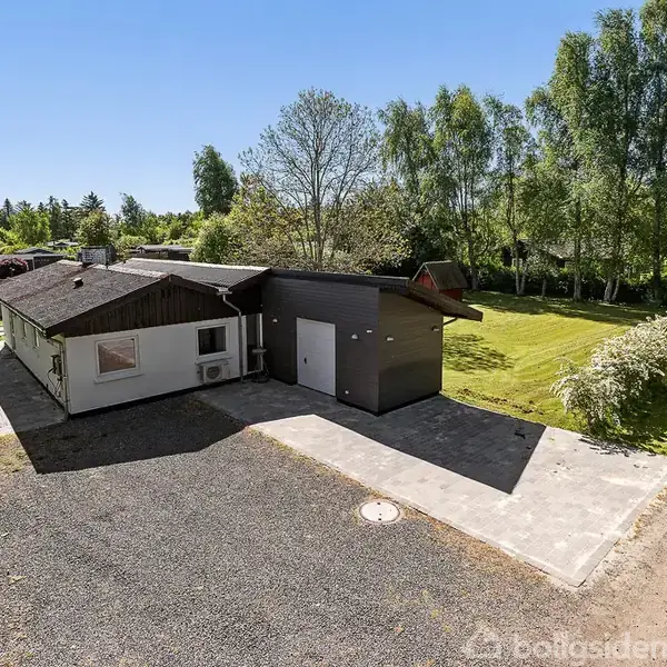 Et hus med fladt tag og garage står på en stor grusbelagt grund. Omkring huset er der en grøn have med træer og buske under en blå himmel.