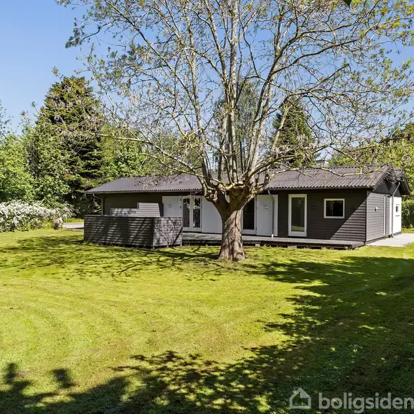 Et mørkt træhus står stille omgivet af en velplejet græsplæne og store træer i en frodig have.