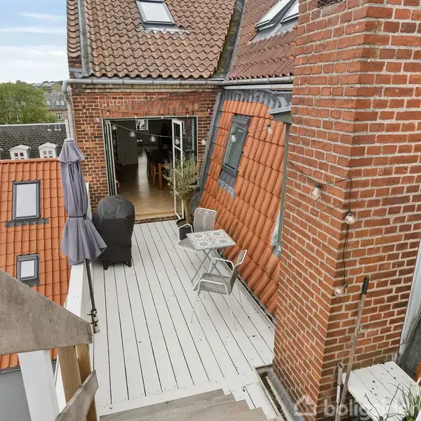 En terrasse med træbord og stole på taget af en bygning. Ved siden af er en grill og en parasol. Trædøre fører ind til en loftslejlighed. Røde tagsten omgiver området.