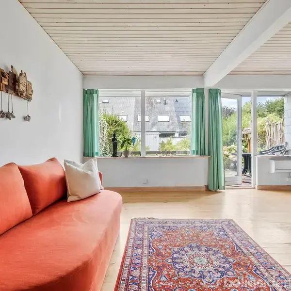 En orange sofa står på et trægulv med et orientalsk tæppe foran. Rummet har store vinduer med grønne gardiner og leder ud til en terrasse med udsigt til haven.