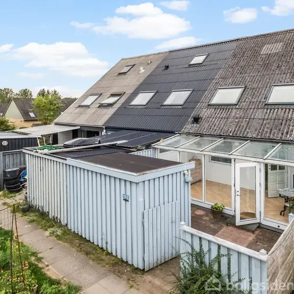 Rækkehus med skrå tagflader og flere ovenlysvinduer, en tilstødende udestue med glasvægge, hvidt skur ved siden af huset; omgivet af blomsterbede og en gangsti.