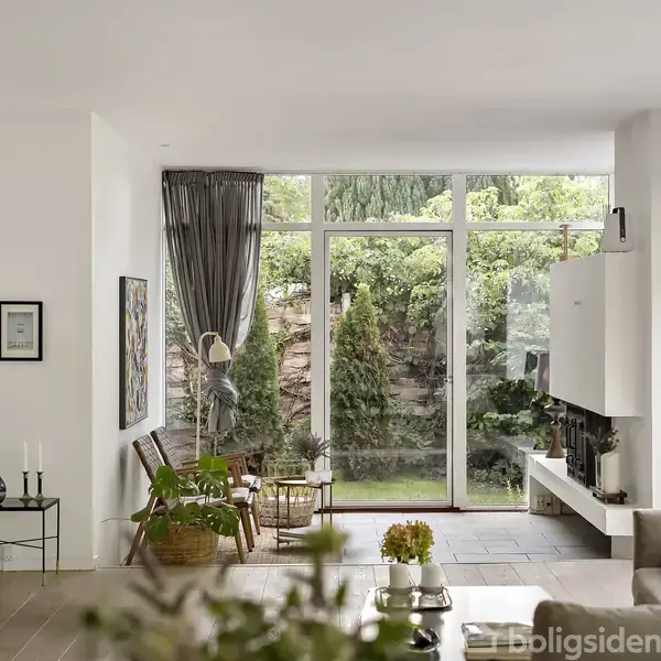 Stue med sofa og planter vender mod stort vindue, der afslører frodig have udenfor. Indretningen inkluderer stearinlys og moderne kunstværker.