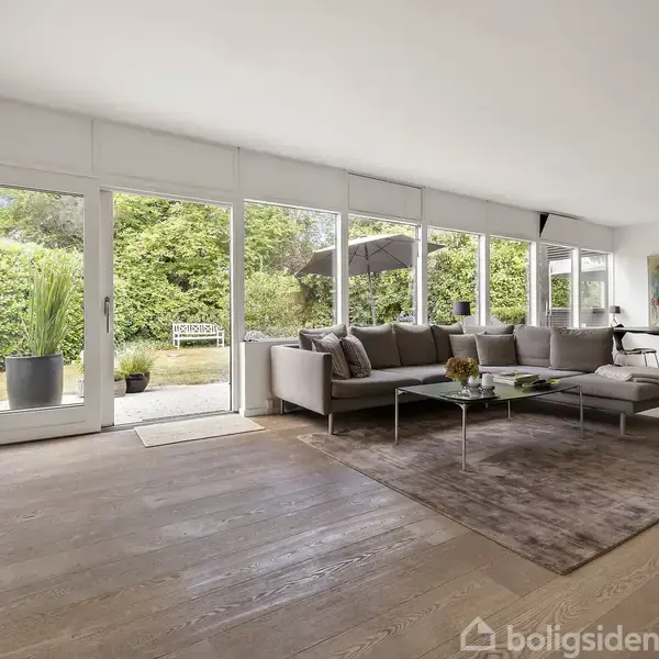 Stue med grå sofa og glasbord står på et tæppe. Store vinduer bagved åbner op til en terrasse med planter. Et spisebord ses i baggrunden.