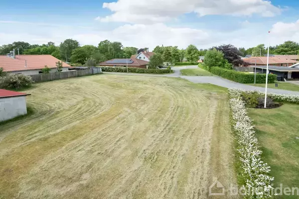 En flad græsplæne omgivet af lave buske ligger i et nabolag med enfamiliehuse og en asfalteret vej i baggrunden under en skyet blå himmel.
