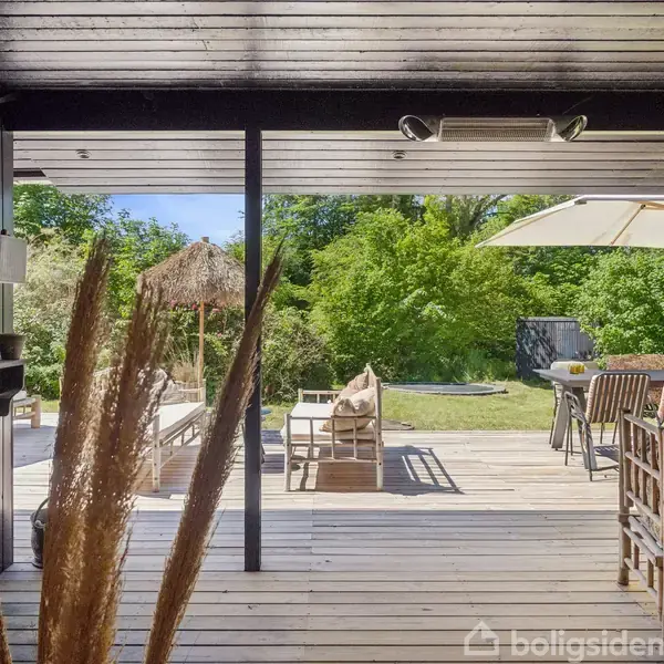 Terrassen har havemøbler, inklusive stole og borde, under en parasol. Omkring terrassen er der frodig vegetation med grønne træer og en blå himmel i baggrunden.