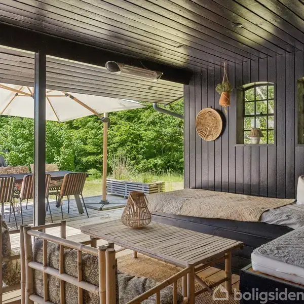 En hyggelig terrasse med træmøbler under et sort, overdækket område. En hvid parasol skygger spisebord og stole. Grønne buske omkranser baggrunden.