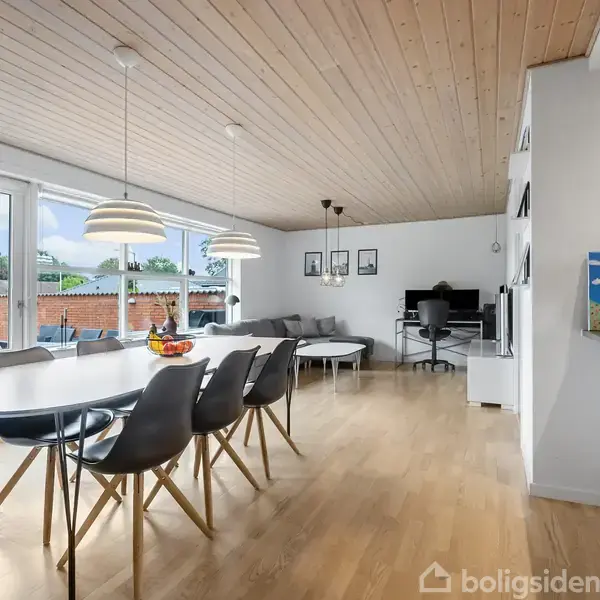 Langbord med sorte stole står i spisestue, hvor lamper hænger over bordet. Store vinduer viser ud til have med murstensvæg. Sofagruppe og reol ses i baggrunden.