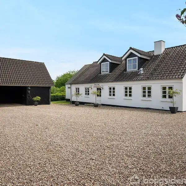 Hvidt hus med mørkt, skråt tag og flere vinduer står på en stor, grusbelagt gårdsplads. Ved siden af huset er der en sort carport med planter. Træer og grønne buske omkranser området.