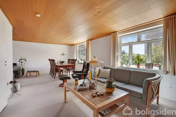 En stue med en grå sofa og sofabord, hvorpå der står dekorationer. Ved siden af er et spisebord med stole. Vinduerne har udsigt til grønne omgivelser.