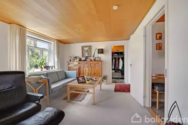 Stue med blå sofa og sort lænestol står omkring et sofabord. Til venstre et vindue med udsigt til grøn natur. Skabsdør åben mod gang med tæppe.