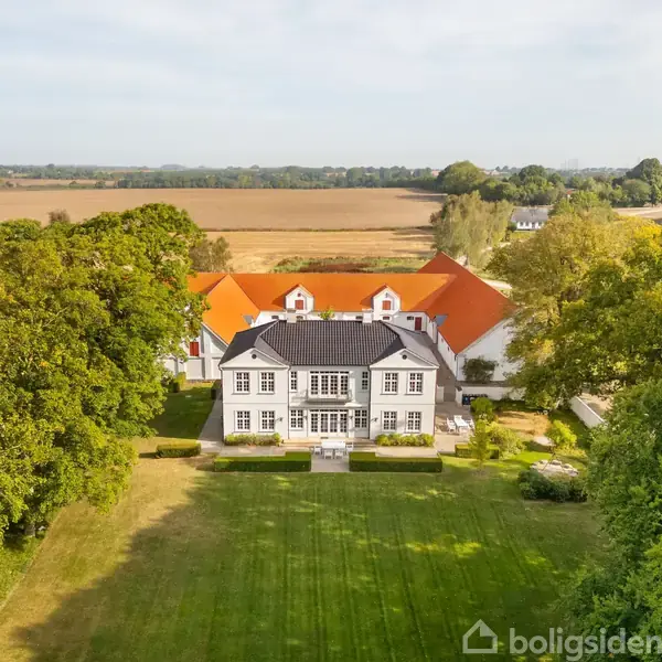 Stort landsted med hvide mure og rødt tag står foran en velplejet, grøn græsplæne. Det er omgivet af træer og åbent landskab i baggrunden.