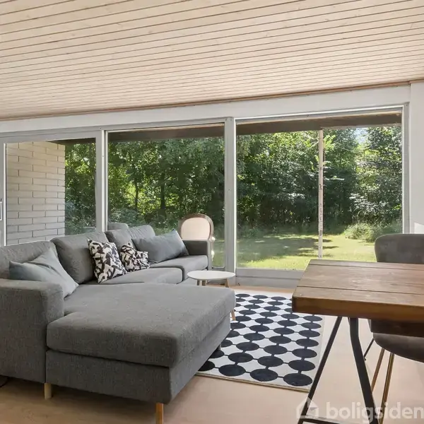 Grå hjørnesofa med pyntepuder står foran store glasvinduer. Udsigt til grøn have. Trægulv og et sort-hvidt tæppe. Træbord med kurv i forgrunden.