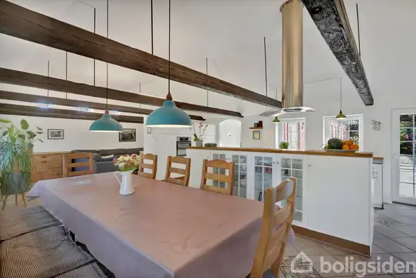 Langbord dækket med lys dug og vase med blomster, omkranset af træstole, under hængende lamper. Stuen har mørke træbjælker i loftet, hvid væg og køkken i baggrunden.