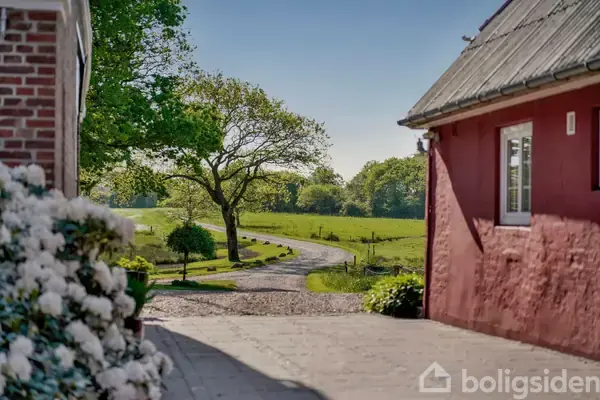 Et hus med rød murstensvæg står i forgrunden. En snoet grusvej fører til en mark med et gammelt træ i baggrunden. Blomster ses til venstre.