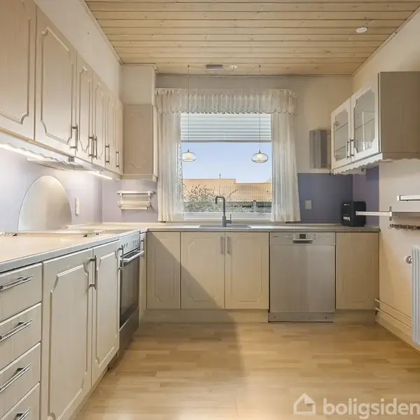 Et lyst køkken med beige skabe og skuffer. En opvaskemaskine og vask er indbygget langs væggen. Et vindue bagved kigger ud på taget af et hus.