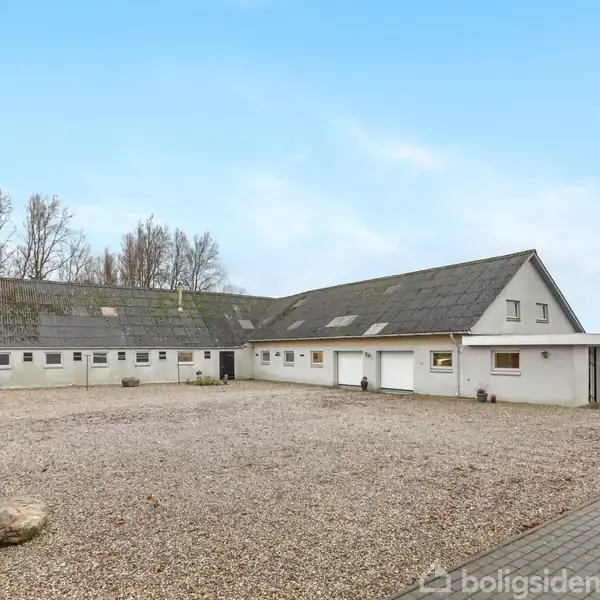 En hvid etplanshus med sort tagstenstag står stille i et åbent landligt område med grusbelagt forplads. Omkring huset er der træer uden blade.