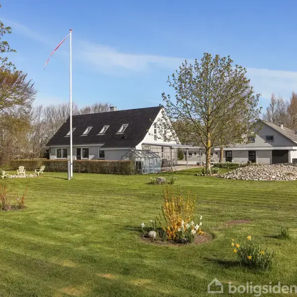 En stor hvid bygning med sort tag står stille i en grøn have. Haven har en flagstang og spredte blomsterbede, omgivet af træer. Teksten "boligsiden" er vandmærket.