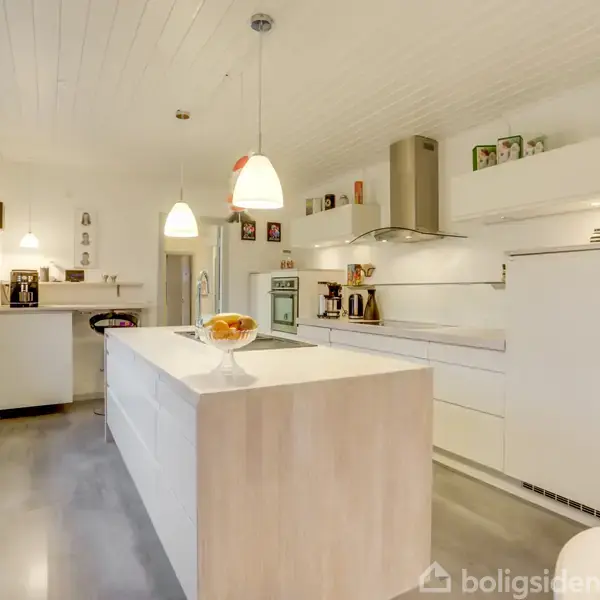 Lyst, moderne køkken med hvidt interiør. Køkkenø med frugtskål. Omgivet af hvide skabe, hængende lamper og køkkenudstyr. Store vinduer slipper lys ind. Ingen tekst fundet.