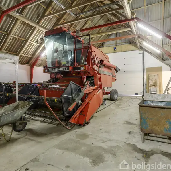Rød mejetærsker står stille i en rummelig lade med synlige bjælker. I forgrunden ses en trillebør, og der er arbejdsborde i baggrunden. Tekst på mejetærsker: "Dronningborg".