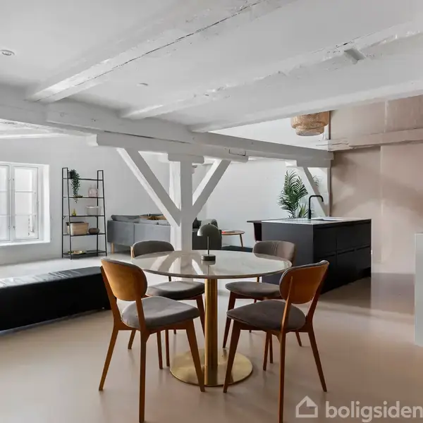 Rundt bord med stole omkring står i et moderne, åbent rum med synlige loftsbjælker. Baggrunden viser et køkken med sorte skabe og en stue med en sofa og reol ved siden af.