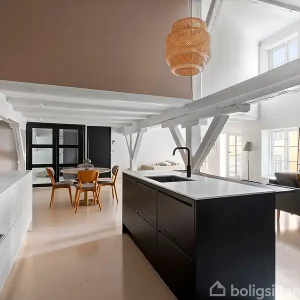 Køkken med sort køkkenø og vandhane, moderne spisebord og stole i baggrunden. Den åbne stue har en grå sofa, reol og store vinduer. Loftet har synlige bjælker.