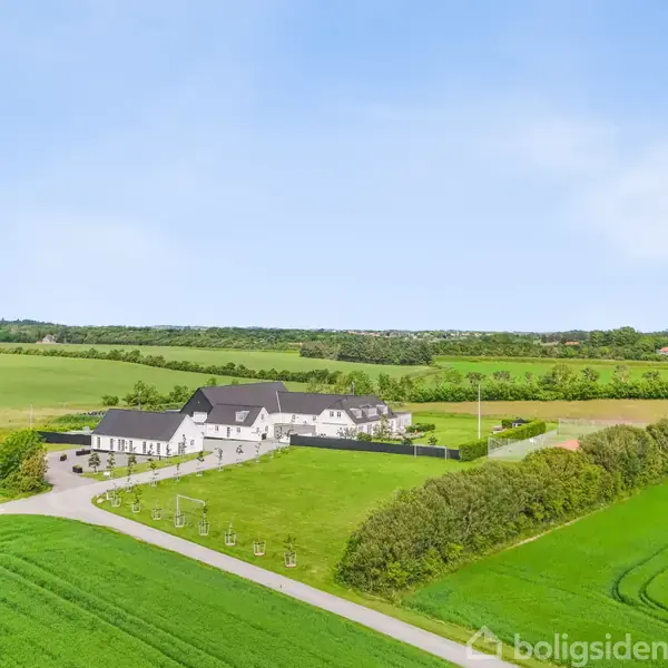 En hvid bygning med sort tag står på et velplejet grønt område med nyplantede træer. Bygningen ligger i et roligt boligområde med asfaltveje og flere lignende huse i baggrunden.