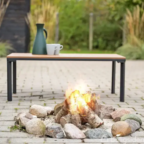 Et bål brænder med sten rundt, placeret på en belagt terrasse. Foran er et lavt bord med en blå kande og en kop. I baggrunden ses en grøn have.