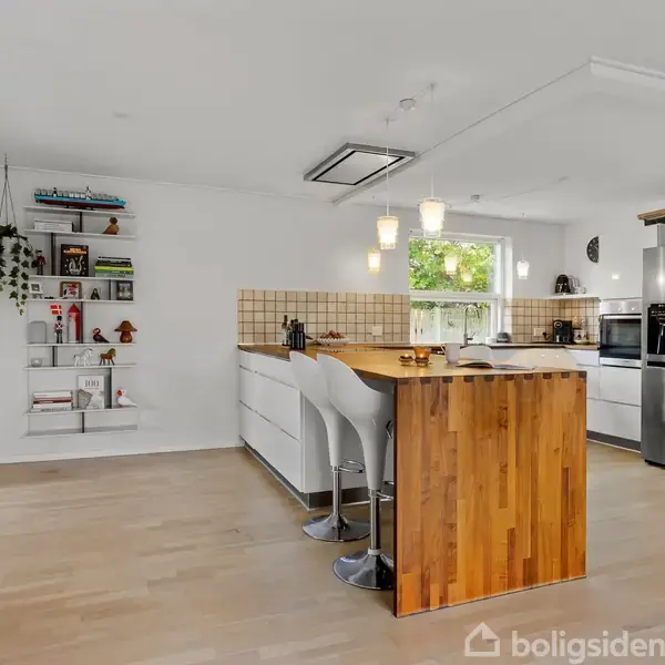 Køkken med træbordplade og tre hvide barstole, lys fliser-splashback, åbne hylder med bøger og planter. Rustfri stål køleskab og bonus-skabe. Lyse krystallysekroner hænger over øen.