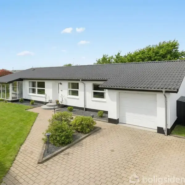 En hvid, enkeltplans villa med sorte tagsten og garage står på en flisebelagt indkørsel omgivet af en velplejet græsplæne og buske i et villakvarter under en klar, blå himmel.