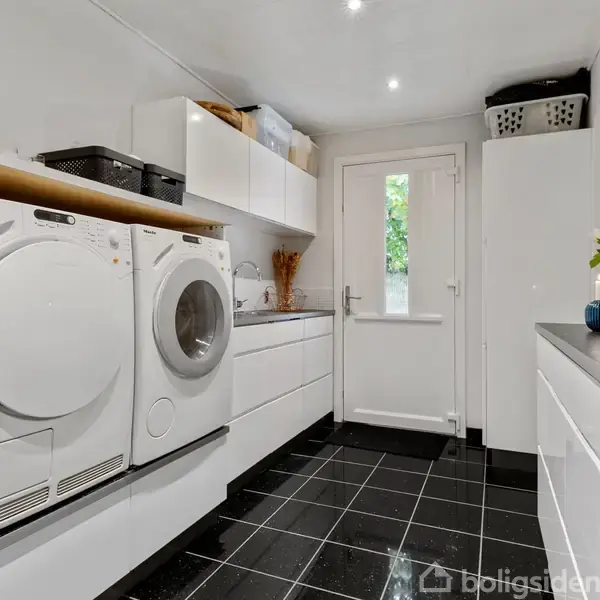 Vaskemaskine og tørretumbler står side om side i et lyst vaskerum med sorte fliser. Hylder er fyldt med kurve, og der er en dekoreret plante på en skænk.