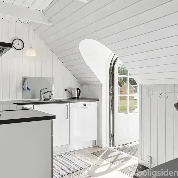 Et lyst køkken med hvide paneler og skrå lofter. Et ur hænger på væggen, mens en dør fører ud til en grøn have. Tekopstilling og kogekande ses på bordpladen.