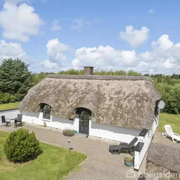 Et bindingsværkshus med stråtag står i en grøn have med træer i baggrunden under en blå himmel med hvide skyer. Foran ses en terrasse med møbler.