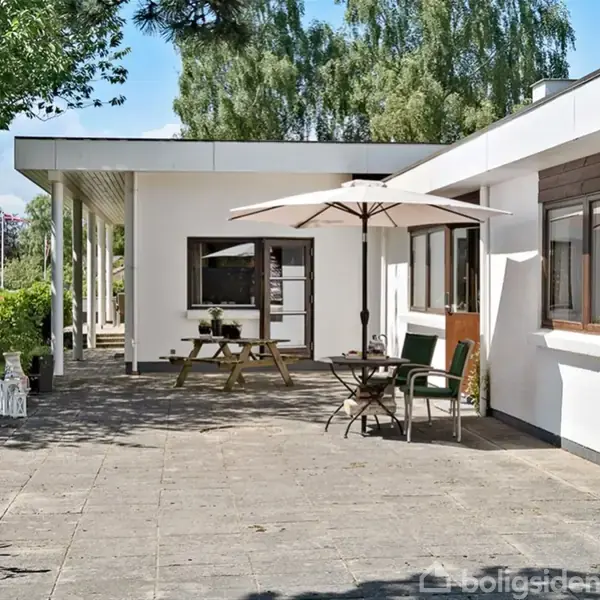En hvid villa har en stor terrasse med fliser; der står et bord med en parasol og stole. Haven omkring er grøn og frodig med træer og buske.