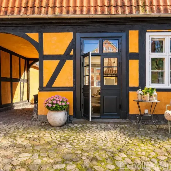 Et bindingsværkshus med sort trækonstruktion og orange vægge, står i gårdhave. En dør og et vindue ses, med blomsterpotte og stol ved siden af, på belagt gårdsplads.