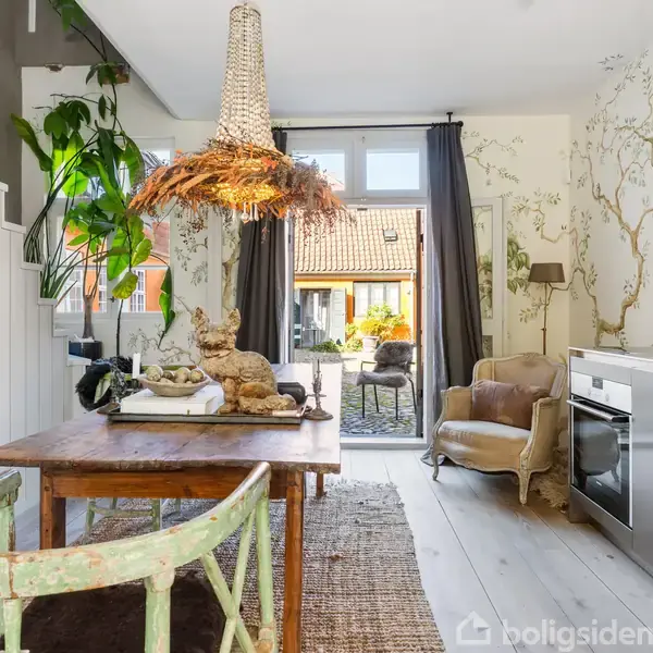 Et spisebord med en rustik lampe ovenover står i et lyst rum. Til venstre er en trappe med planter, og til højre ses en ovn og en lænestol. Væggene har naturinspireret tapet med træer, og glasdøre i baggrunden fører til en gårdhave.
