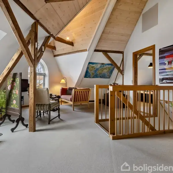Loftstue med træbjælker og skråt loft indeholder en gyngehest, lænestole, kunst på væggene og planter. Store vinduer giver lys til rummet, som er indrettet til afslapning.