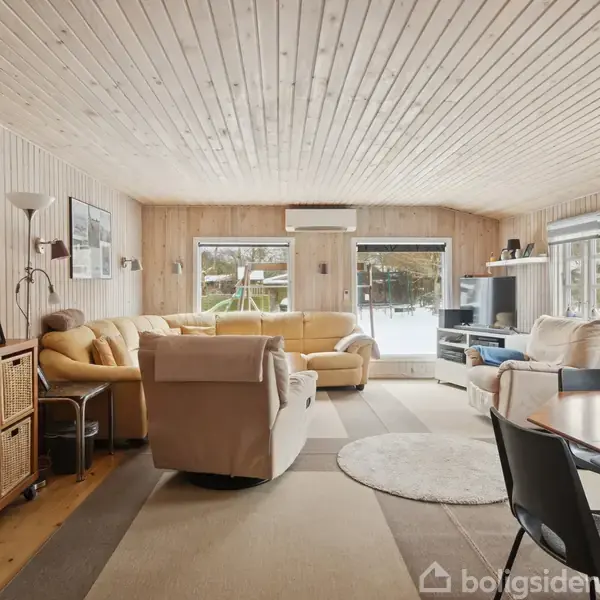 Stue med lyse trævægge, beige sofa og lænestole på tæppe, udsigt til sneklædt have gennem store vinduer, spisebord med stole i forgrunden, og reolsystem med kurve.