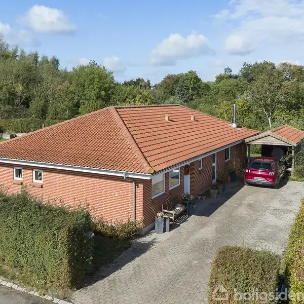 En murstensvilla med rødt tegltag står parkeret med en bil i carporten. Omkring huset er der en brostensbelagt indkørsel og hække, omgivet af et grønt skovområde.