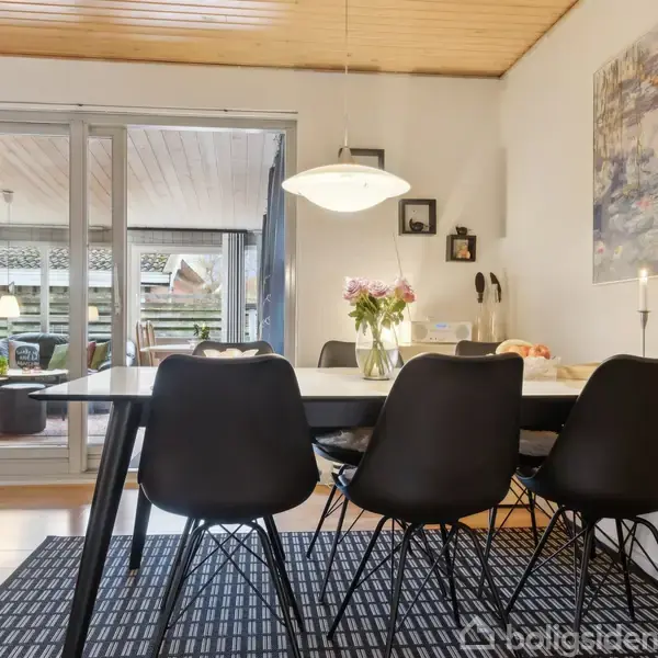 Spisebord med fem sorte stole står i en veloplyst spisestue. Bordet er dekoreret med en vase med blomster og stearinlys. Rummet har lys trægulv og åbner op til stue.