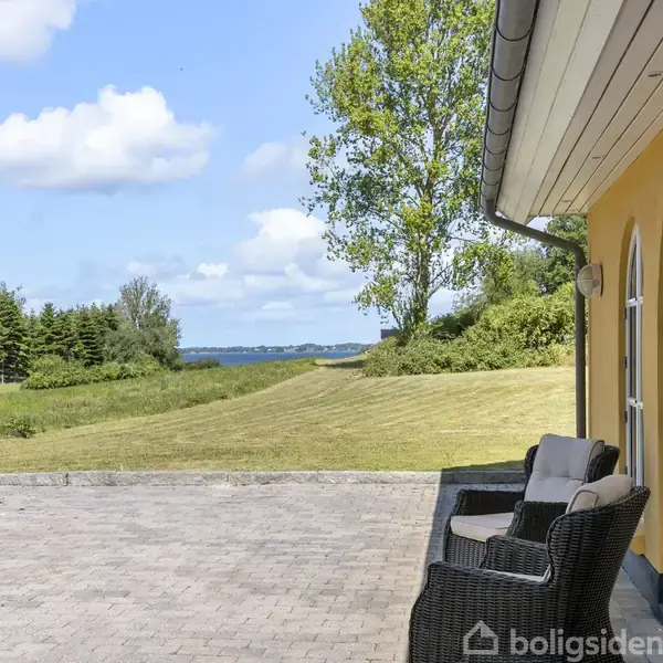 Terrasse med flettestole står foran et hus med buede vinduer. Haven har en græsplæne og træer, og i baggrunden ses havet og en skov. Skyer pryder himlen.