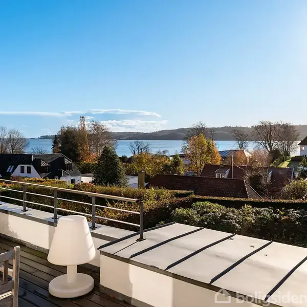 Terrasse med hvide møbler og lille lampe, der står stille under en klar blå himmel. Udsigt over en sø med omgivende huse og træer i efterårsfarver.